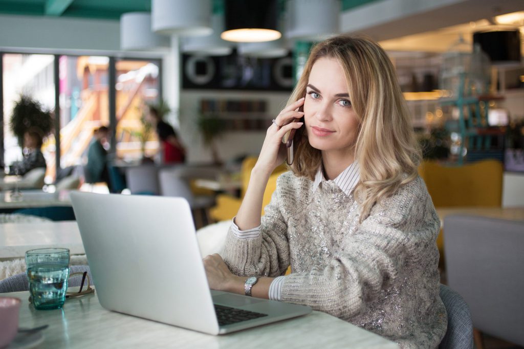 Portrait of beautiful female manager talking on phone. Confident young business lady calling colleague from cafe. She sitting at table with open laptop and looking at camera. Businesswoman concept